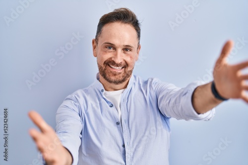 Middle age caucasian man standing over blue background looking at the camera smiling with open arms for hug. cheerful expression embracing happiness.