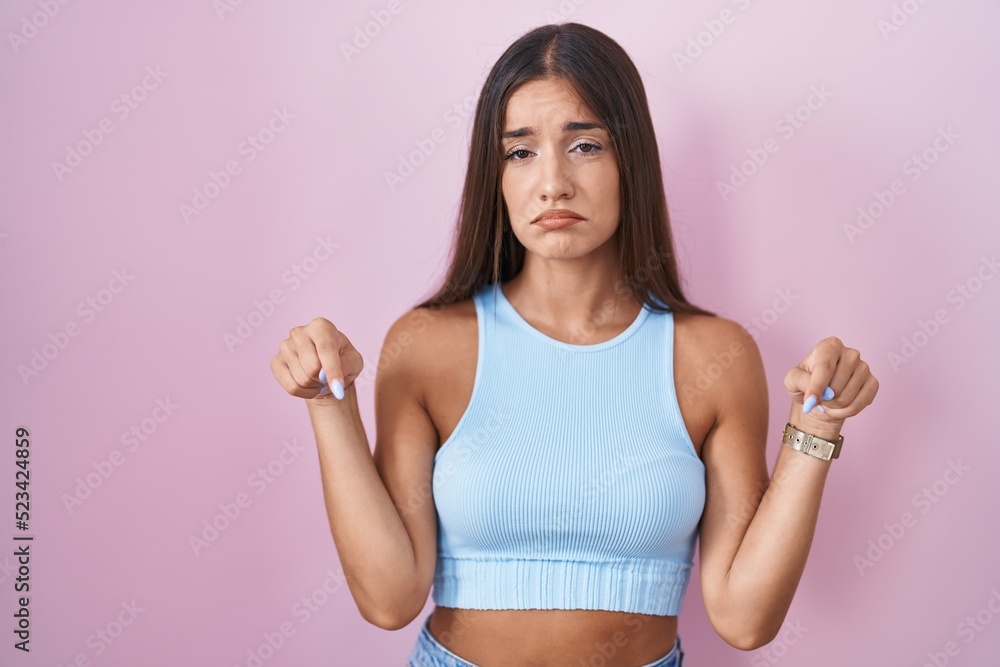 Young brunette woman standing over pink background pointing down looking sad and upset, indicating direction with fingers, unhappy and depressed. Young brunette woman standing over pink background pointing down looking sad and upset, indicating direction with fingers, unhappy and depressed.