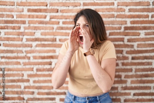 Young brunette woman standing over bricks wall shouting angry out loud with hands over mouth