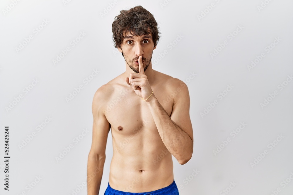 Fototapeta premium Young hispanic man standing shirtless over isolated, background asking to be quiet with finger on lips. silence and secret concept.