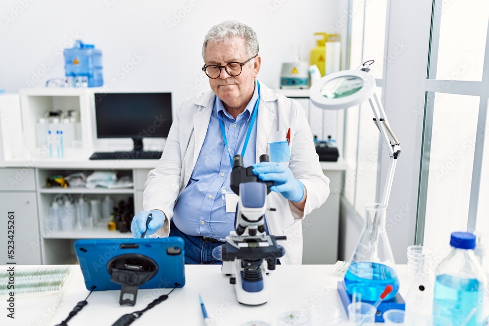 Middle age grey-haired man wearing scientist uniform using microscope and touchpad at laboratory Middle age grey-haired man wearing scientist uniform using microscope and touchpad at laboratory