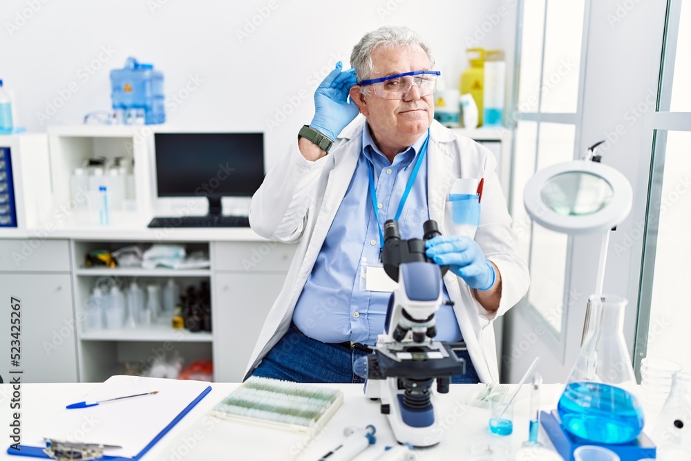 Senior caucasian man working at scientist laboratory smiling with hand over ear listening an hearing to rumor or gossip. deafness concept. Senior caucasian man working at scientist laboratory smiling with hand over ear listening an hearing to rumor or gossip. deafness concept.
