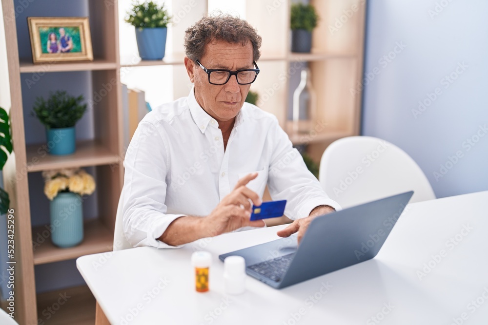 Middle age man using laptop and credit card sitting on table with pills bottles at home Middle age man using laptop and credit card sitting on table with pills bottles at home