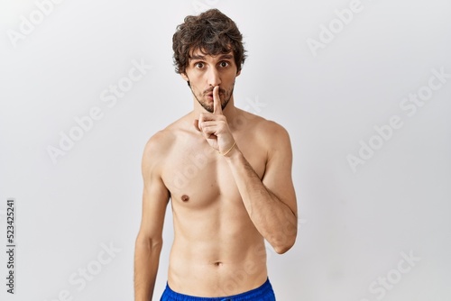 Young hispanic man standing shirtless over isolated, background asking to be quiet with finger on lips. silence and secret concept.