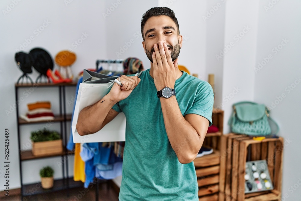 Young handsome man with beard holding shopping bags at retail shop laughing and embarrassed giggle covering mouth with hands, gossip and scandal concept Young handsome man with beard holding shopping bags at retail shop laughing and embarrassed giggle covering mouth with hands, gossip and scandal concept