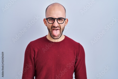 Young bald man with beard standing over white background wearing glasses sticking tongue out happy with funny expression. emotion concept. © Krakenimages.com