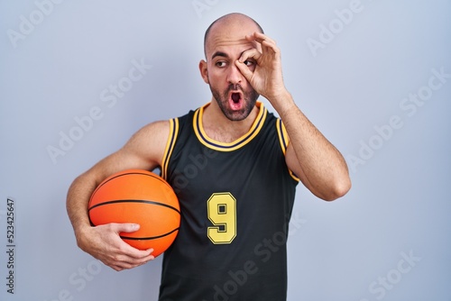 Young bald man with beard wearing basketball uniform holding ball doing ok gesture shocked with surprised face, eye looking through fingers. unbelieving expression.
