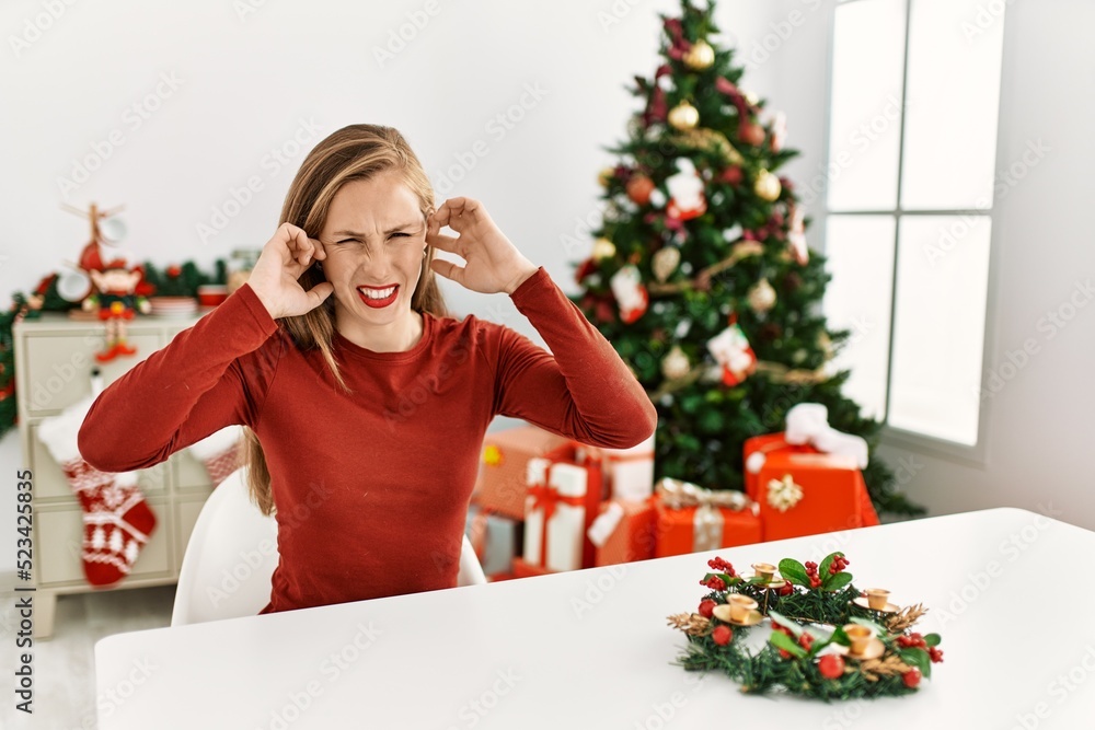 Caucasian young blonde woman sitting on the table by christmas tree covering ears with fingers with annoyed expression for the noise of loud music. deaf concept. Caucasian young blonde woman sitting on the table by christmas tree covering ears with fingers with annoyed expression for the noise of loud music. deaf concept.