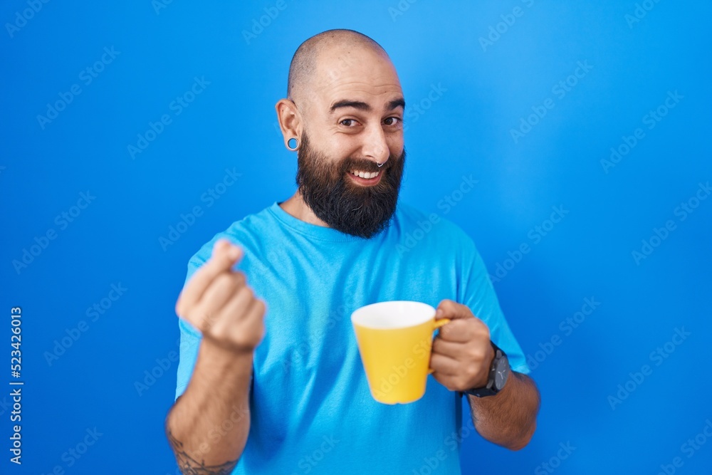 Young hispanic man with beard and tattoos drinking a cup of coffee doing money gesture with hands, asking for salary payment, millionaire business Young hispanic man with beard and tattoos drinking a cup of coffee doing money gesture with hands, asking for salary payment, millionaire business