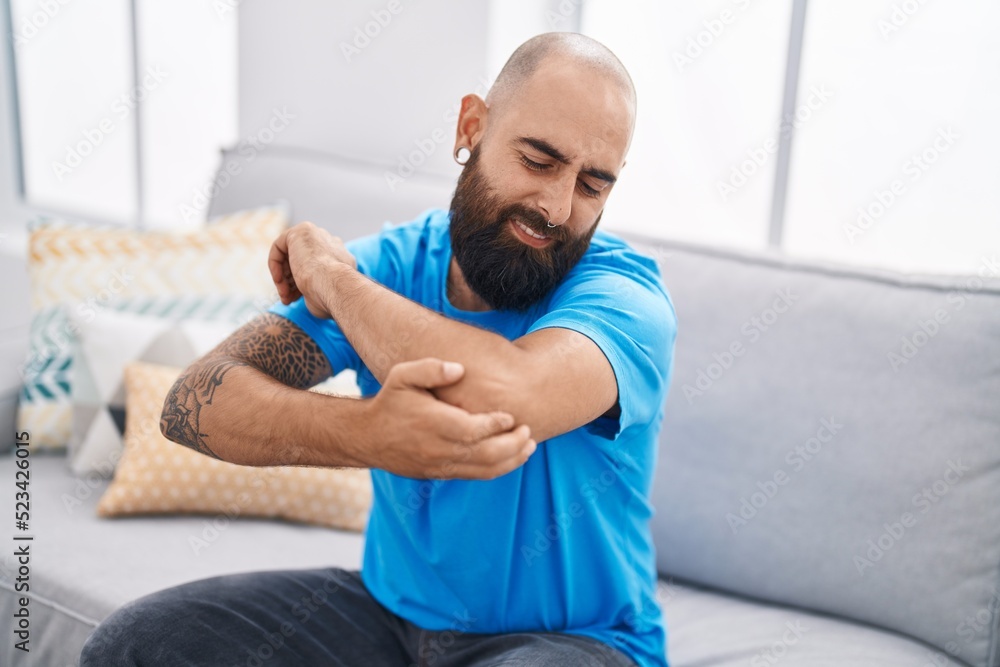 Young bald man sitting on sofa suffering for elbow pain at home Young bald man sitting on sofa suffering for elbow pain at home
