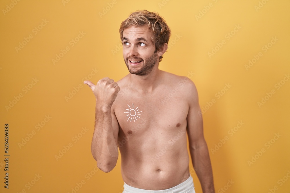 Caucasian man standing shirtless wearing sun screen smiling with happy face looking and pointing to the side with thumb up.