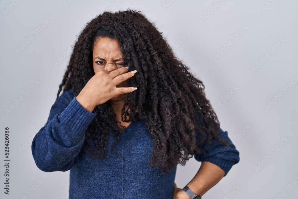 Plus size hispanic woman standing over white background smelling something stinky and disgusting, intolerable smell, holding breath with fingers on nose. bad smell Plus size hispanic woman standing over white background smelling something stinky and disgusting, intolerable smell, holding breath with fingers on nose. bad smell