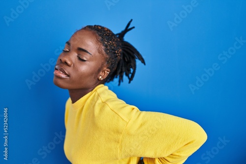 Beautiful black woman standing over blue background suffering of backache, touching back with hand, muscular pain