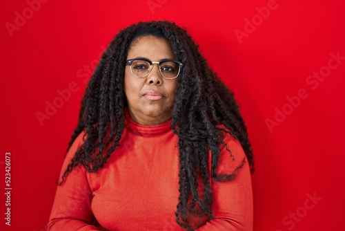 Plus size hispanic woman standing over red background relaxed with serious expression on face. simple and natural looking at the camera. © Krakenimages.com
