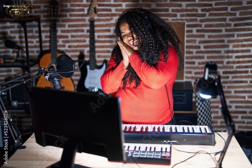 Plus size hispanic woman playing piano at music studio sleeping tired dreaming and posing with hands together while smiling with closed eyes.