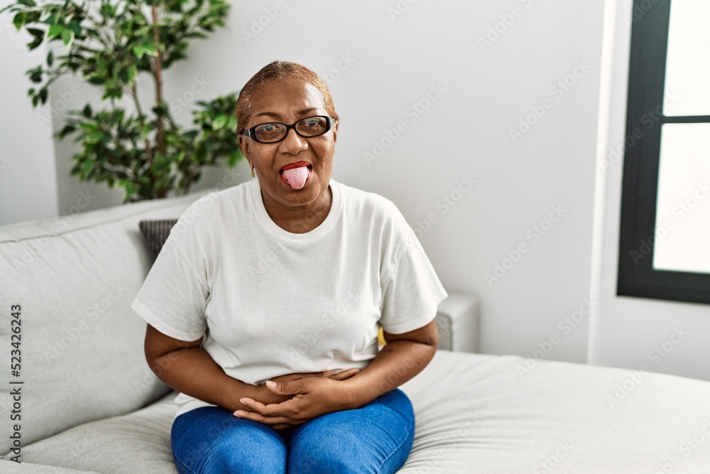 Mature hispanic woman sitting on the sofa at home sticking tongue out happy with funny expression. emotion concept.