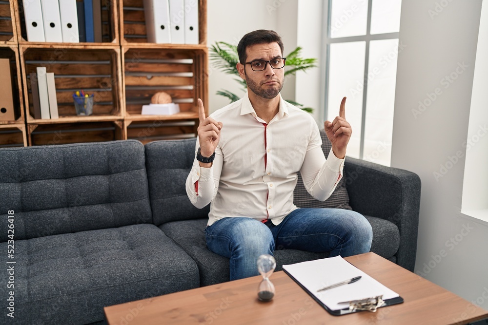 Young hispanic man with beard working at consultation office pointing up looking sad and upset, indicating direction with fingers, unhappy and depressed. Young hispanic man with beard working at consultation office pointing up looking sad and upset, indicating direction with fingers, unhappy and depressed.