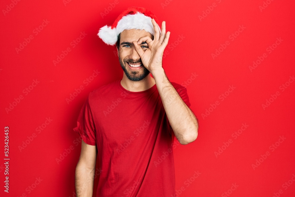 Young hispanic man wearing christmas hat doing ok gesture with hand smiling, eye looking through fingers with happy face. Young hispanic man wearing christmas hat doing ok gesture with hand smiling, eye looking through fingers with happy face.