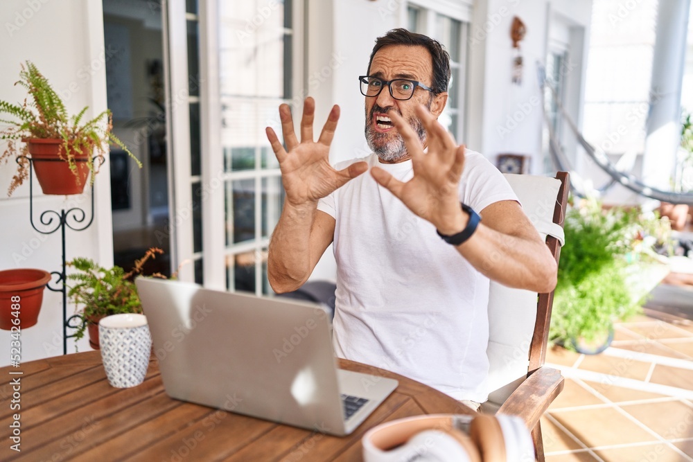 Middle age man using computer laptop at home afraid and terrified with fear expression stop gesture with hands, shouting in shock. panic concept. Middle age man using computer laptop at home afraid and terrified with fear expression stop gesture with hands, shouting in shock. panic concept.