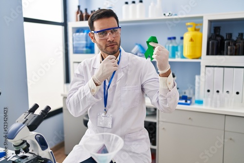 Young hispanic man with beard working at scientist laboratory holding green ribbon serious face thinking about question with hand on chin  thoughtful about confusing idea