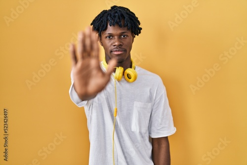 Young african man with dreadlocks standing over yellow background doing stop sing with palm of the hand. warning expression with negative and serious gesture on the face.