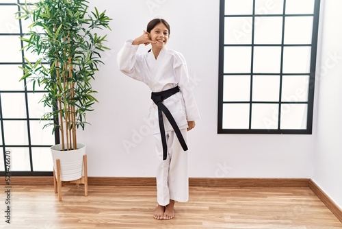 Young hispanic girl wearing karate kimono and black belt smiling doing phone gesture with hand and fingers like talking on the telephone. communicating concepts.