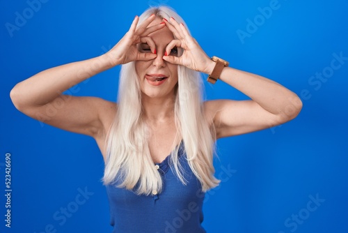 Caucasian woman standing over blue background doing ok gesture like binoculars sticking tongue out, eyes looking through fingers. crazy expression.