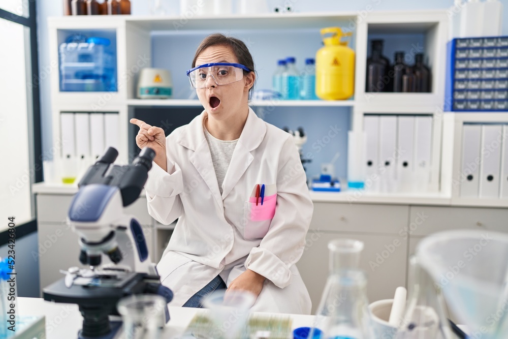 Hispanic girl with down syndrome working at scientist laboratory surprised pointing with finger to the side, open mouth amazed expression. Hispanic girl with down syndrome working at scientist laboratory surprised pointing with finger to the side, open mouth amazed expression.