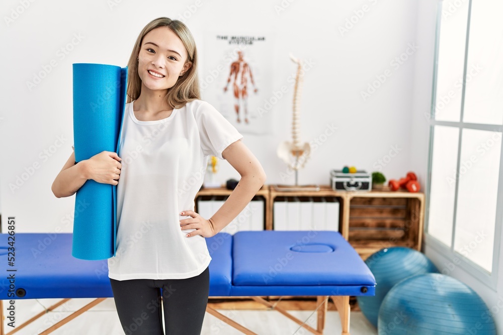 Young chinese woman smiling confident holding yoga mat at clinic