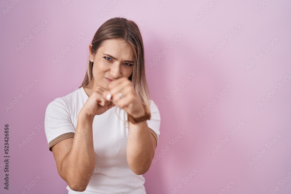 Blonde caucasian woman standing over pink background punching fist to fight, aggressive and angry attack, threat and violence Blonde caucasian woman standing over pink background punching fist to fight, aggressive and angry attack, threat and violence