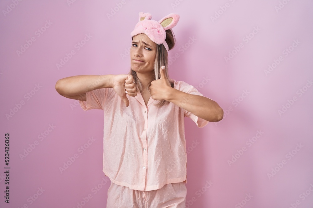 Blonde caucasian woman wearing sleep mask and pajama doing thumbs up and down, disagreement and agreement expression. crazy conflict