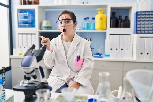 Hispanic girl with down syndrome working at scientist laboratory surprised pointing with finger to the side  open mouth amazed expression.