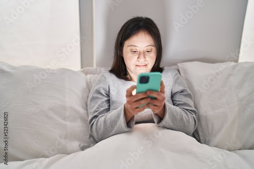 Young woman with down syndrome using smartphone sitting on bed at bedroom