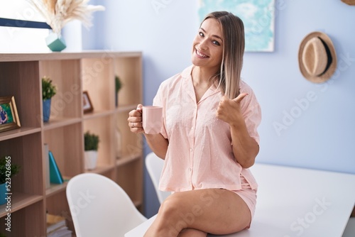 Blonde caucasian woman drinking a cup coffee wearing pajama smiling happy and positive  thumb up doing excellent and approval sign