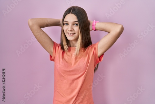 Teenager girl standing over pink background relaxing and stretching, arms and hands behind head and neck smiling happy