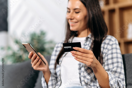 Defocused caucasian pretty girl, holding a smartphone and a credit card in her hands, makes online purchases in online stores, orders home delivery, smiling happily. Online shopping concept