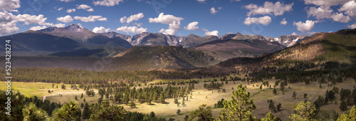 Panoramic landscape scene of a valley in Rocky Mountain National Park, Colorado during the summer. There are white fluffy clouds in the sky and the sun is shining onto the valley floor. 