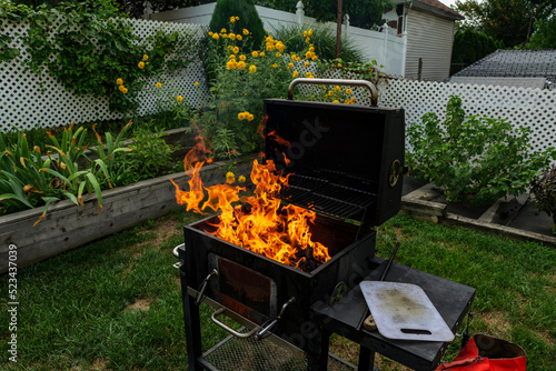 Brightly burning in metal box logs firewood for barbecue outdoor on sunny day. Orange high flame and white smoke on blurred blue sky and green grass background. Camping, safety and tourism concept