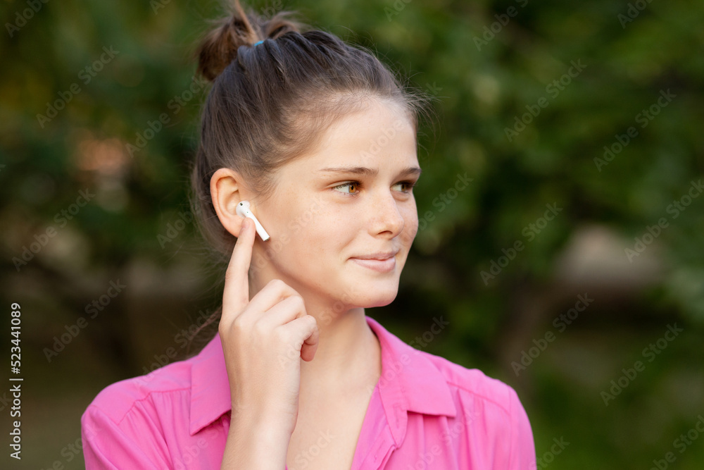 Close up portrait of lovely girl in pink listening to music through wireless earphones outdoors. Young girl smiling happy. Airpods. Close up portrait of lovely girl in pink listening to music through wireless earphones outdoors. Young girl smiling happy. Airpods.