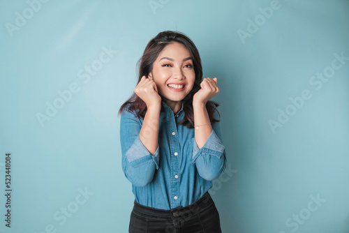 A portrait of a cute Asian woman wearing a blue shirt and feeling excited and isolated by a blue background