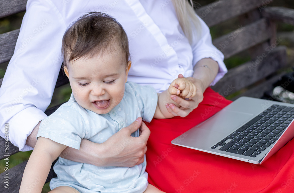 young mother using laptop and sitting with little baby on park bench,outside.freelancer job during maternity concept.business lady working,typing on keyboard.baby make different faces multitasking mom young mother using laptop and sitting with little baby on park bench,outside.freelancer job during maternity concept.business lady working,typing on keyboard.baby make different faces multitasking mom