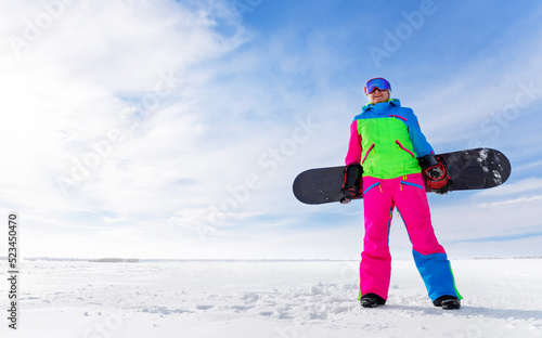 Young girl holds a snowboard in her hands on the background of a winter landscape. Woman in ski goggles and overalls with her board goes in for sports on a sunny frosty day.