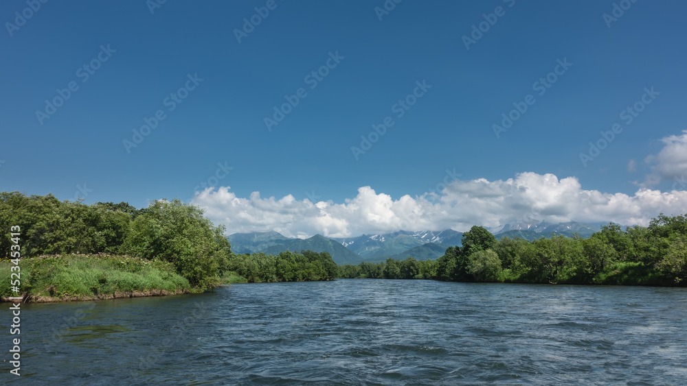 The bed of the blue river bends between the banks overgrown with lush green vegetation. A mountain range in the distance, against the sky and clouds. Kamchatka. River Bystraya