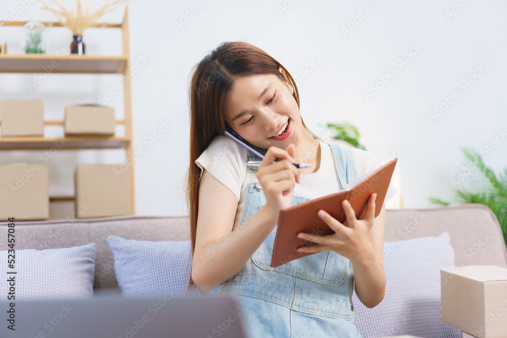 Online merchant concept, Female entrepreneur taking notes in notebook while talking on smartphone Online merchant concept, Female entrepreneur taking notes in notebook while talking on smartphone