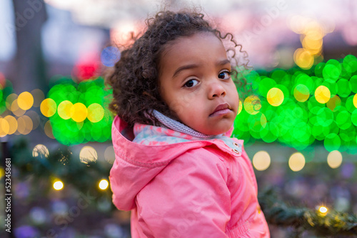 Adorable mixed race girl looks at camera in front of Christmas lights bokeh