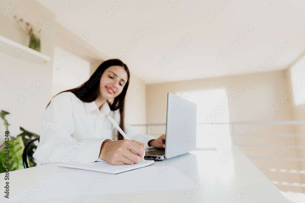 Caucasian young business woman sitting at desk, reading from computer, taking notes at her white notebook. Student working from home during online work meeting or webinar. Happy smiling lady at office Caucasian young business woman sitting at desk, reading from computer, taking notes at her white notebook. Student working from home during online work meeting or webinar. Happy smiling lady at office