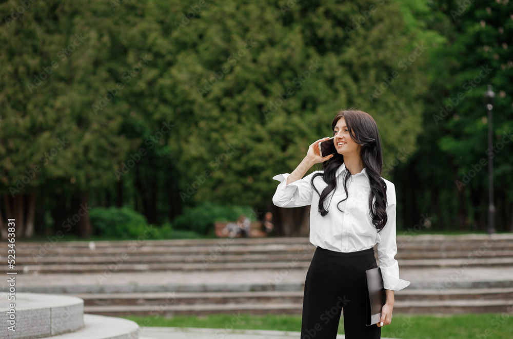 A pretty woman in a white shirt stands in the park with a folder in hand talking on a smartphone. A pretty woman in a white shirt stands in the park with a folder in hand talking on a smartphone.