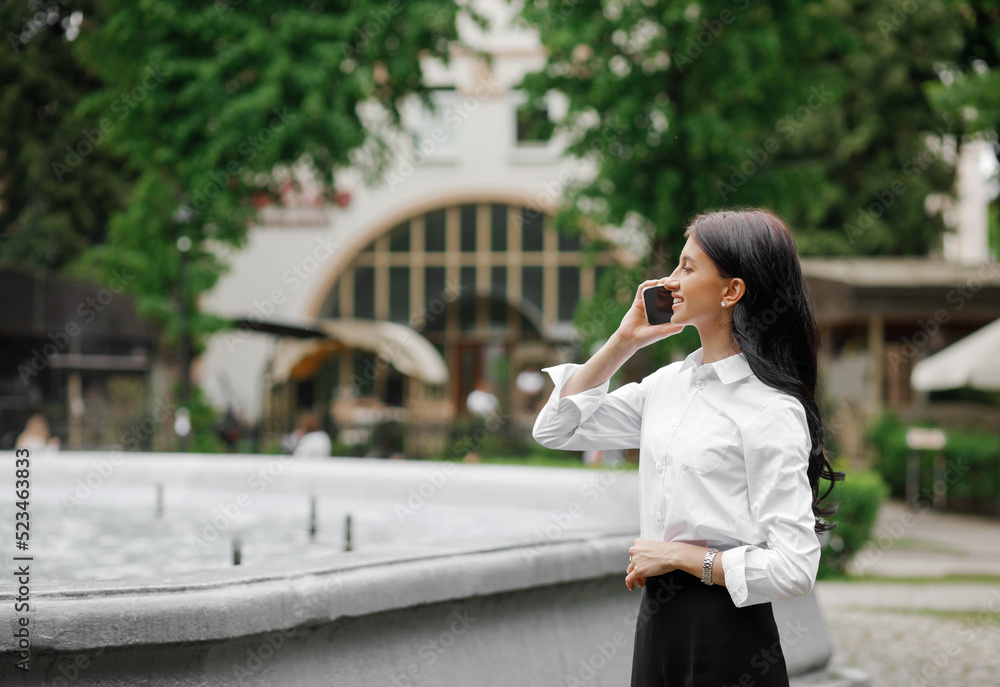 Fototapeta premium Beautiful, caucasian girl talking on the phone on the background of a fountain in the park. Freelance concept.