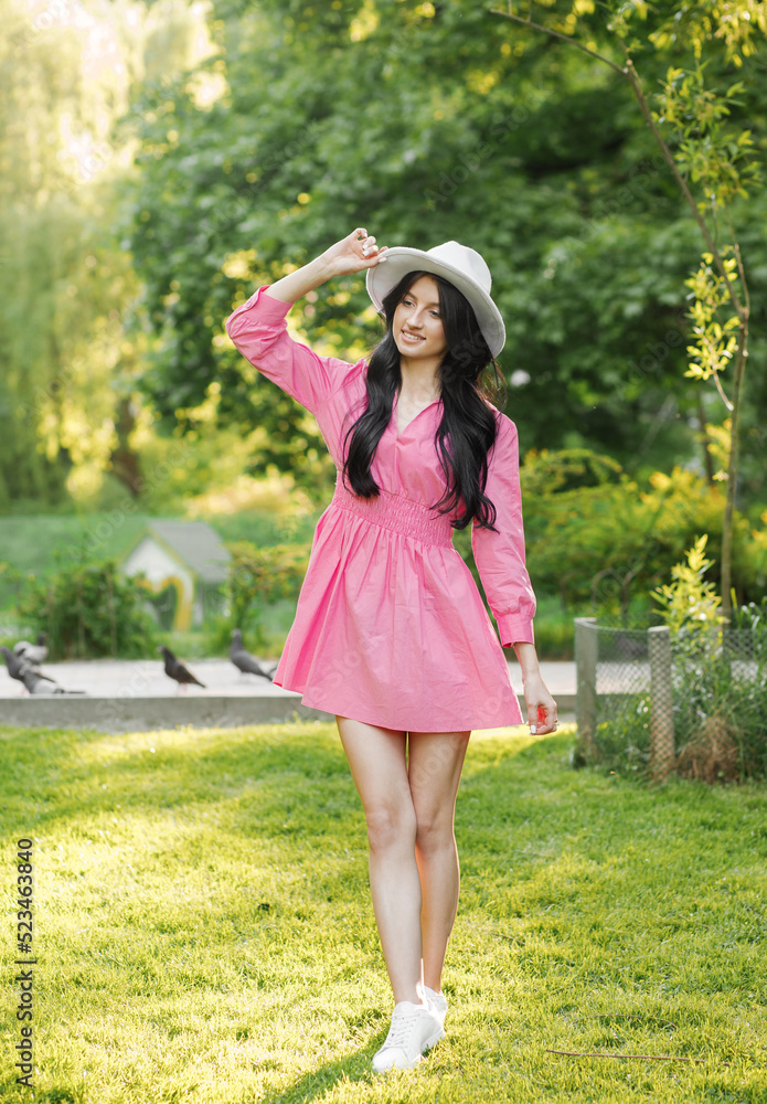Photo of a cute lady in a pink dress and white hat smiling while walking in the park