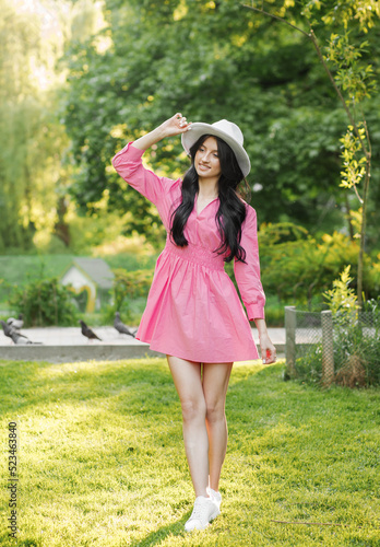 Photo of a cute lady in a pink dress and white hat smiling while walking in the park © Yuliia